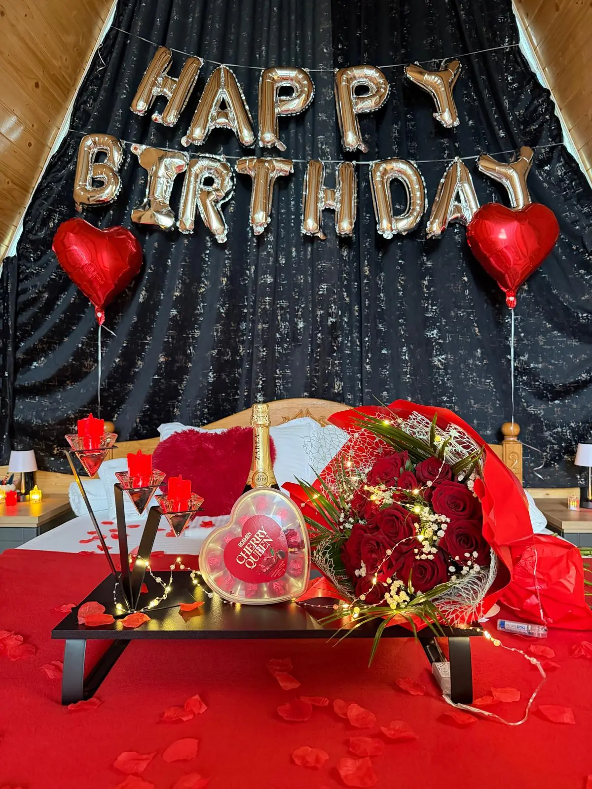 Baloane 'Happy Birthday' și inimi roșii deasupra unui pat decorat cu trandafiri, șampanie și bomboane